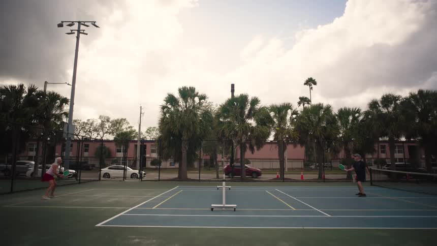 Two older people play pickleball on a tennis court while traffic passes on a nearby street on a cloudy summer day
