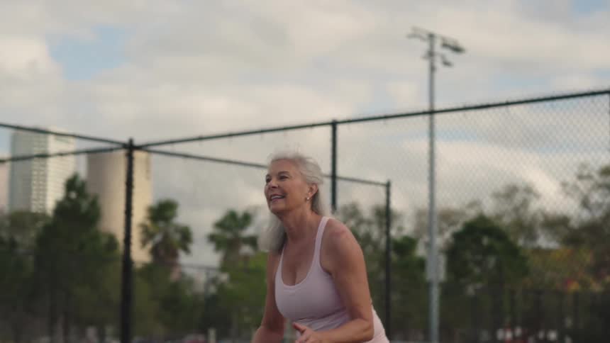 A smiling senior woman energetically hits a pickleballtennis ball upwards while playing on an outdoor court with city buildings visible in the background