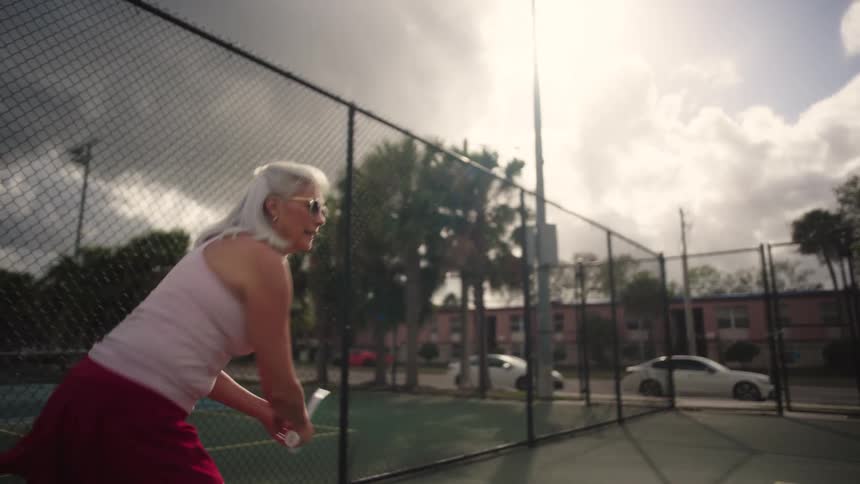 Elderly woman plays pickleball outdoors on a court surrounded by fences
