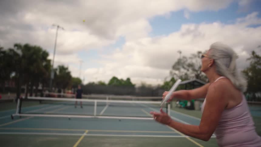 Seen from behind an active senior woman follows through on a backhand shot while playing pickleball or tennis on an outdoor court under a partly cloudy sky