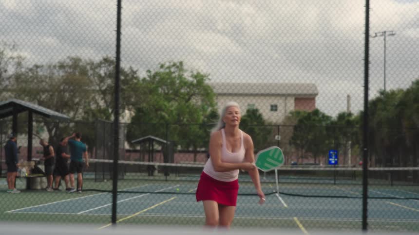 An older woman plays pickleball on a court on a cloudy summer day