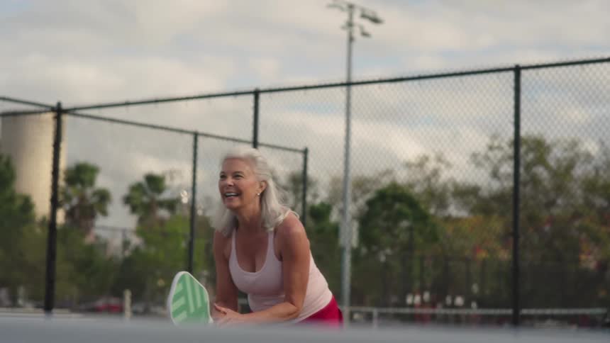 A smiling active senior woman hits a return shot while playing tennis on an outdoor court near city buildings
