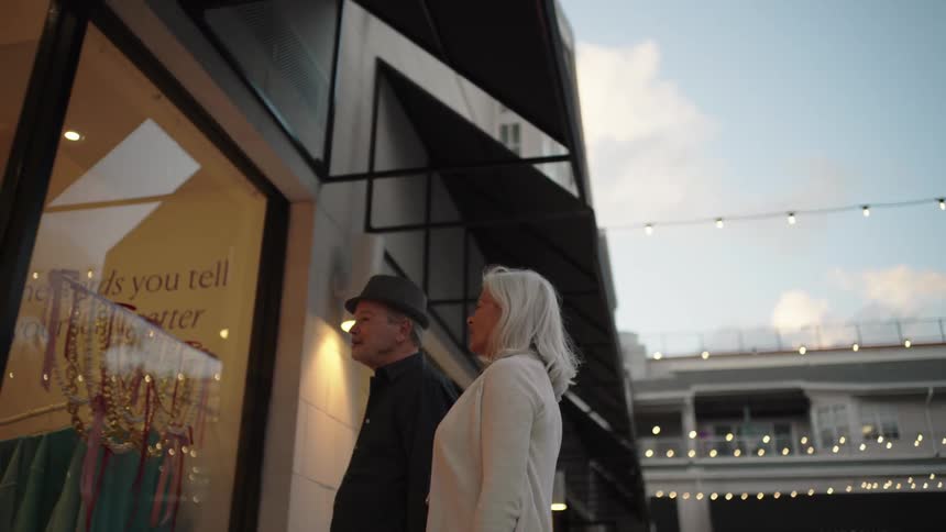 A senior couple window shops along an outdoor promenade at dusk pointing and talking as they look at a store display beneath string lights
