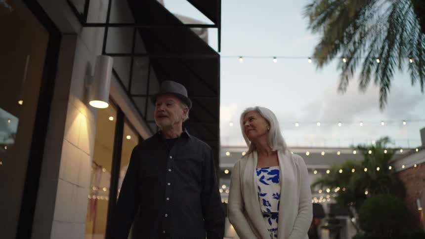 Mature couple walk side by side in a shopping area lit by decorative lights at dusk