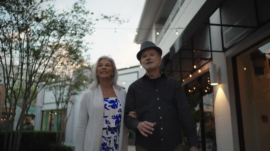A senior couple walks arminarm talking and gesturing while strolling through an outdoor shopping area lined with trees and lit storefronts at dusk