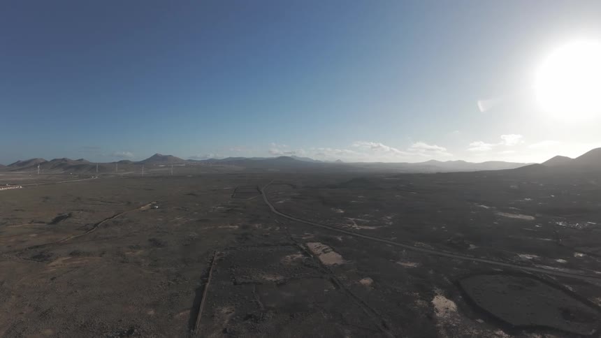 A high aerial view reveals a vast arid landscape stretching towards distant mountains and wind turbines under a bright clear blue sky