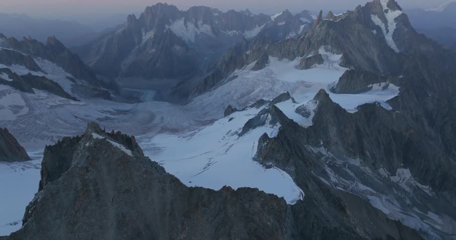 An aerial view reveals a vast rugged mountain range with jagged peaks covered in snow and glaciers illuminated by the soft light of dawn or dusk