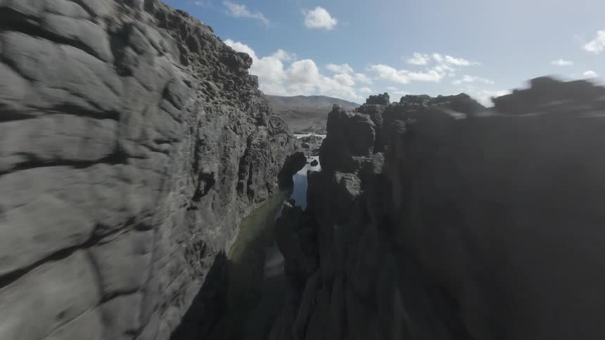 A rugged rocky coastline with crashing waves under a partly cloudy sky extending towards distant hills