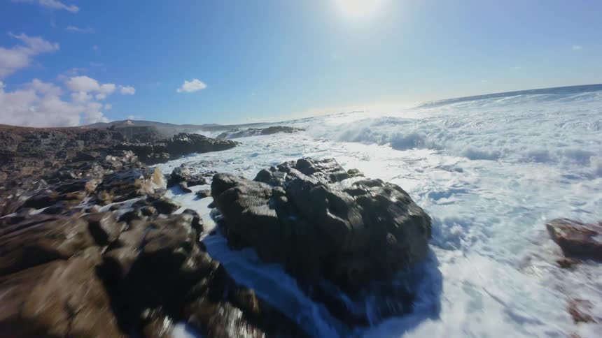 Aerial of ocean waves crashing into a rocky coastline on a mostly sunny day