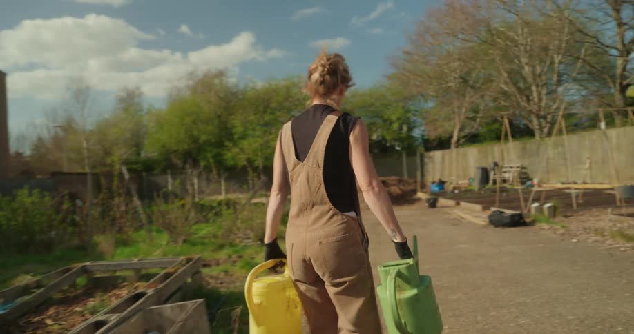 Female gardener working in the area behind a row of apartment buildings uses watering cans to water the plants in a garden bed on a cloudy day