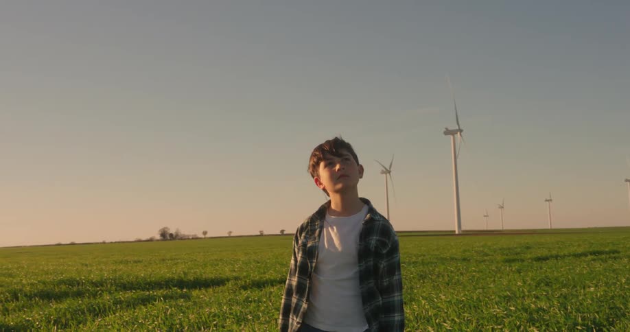 A boy stands in a green field with several wind turbines scattered across the landscape under a clear blue sky
