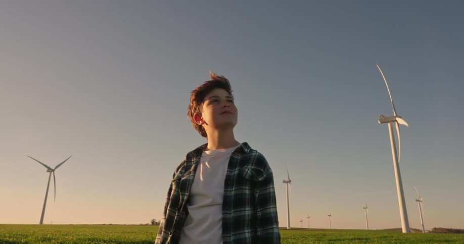 A teenage boy stands outdoors and looks up at a windmill on a windy summer day