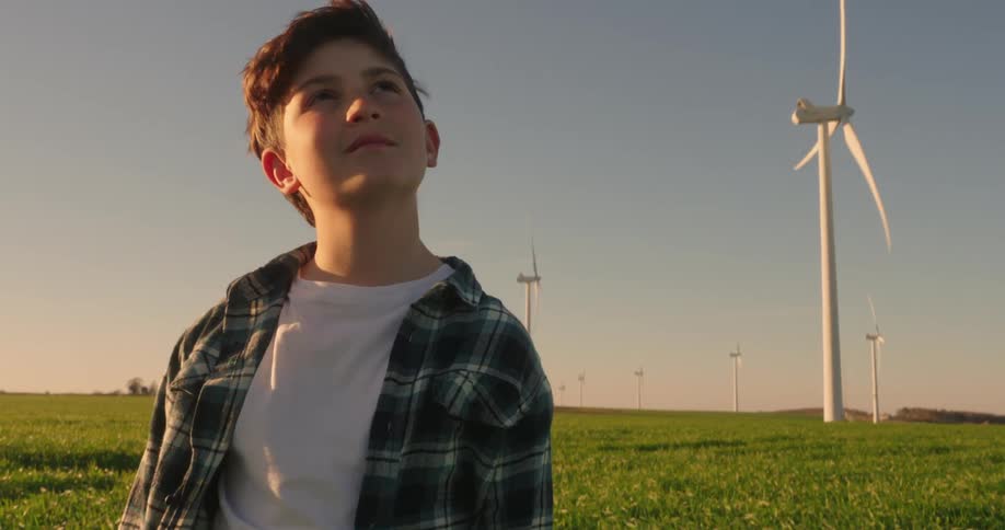 A young boy wearing a plaid shirt over a white tee gazes upwards at a towering wind turbine against a warm toned sky