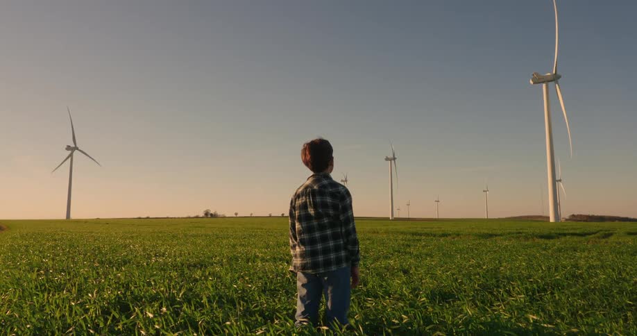 A boy stands outdoors on a wind farm and looks up at a windmill on a windy summer day