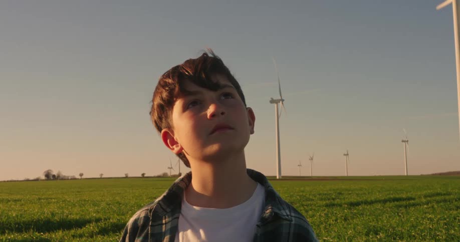 A boy stands in a green field with several wind turbines scattered across the landscape under a clear blue sky