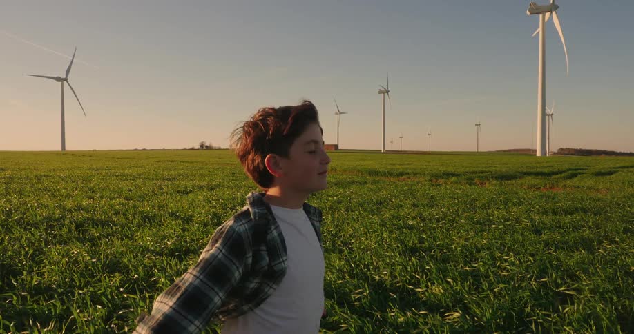 With eyes closed and arms outstretched to the wind a young person stands in a golden field beside wind turbines