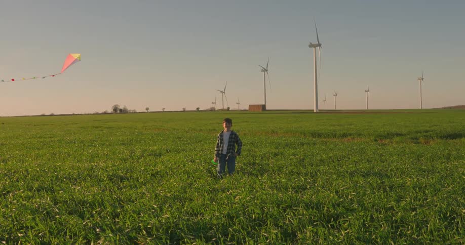 Child in green field watches a kite in the sky at dusk surrounded by wind turbines in the background