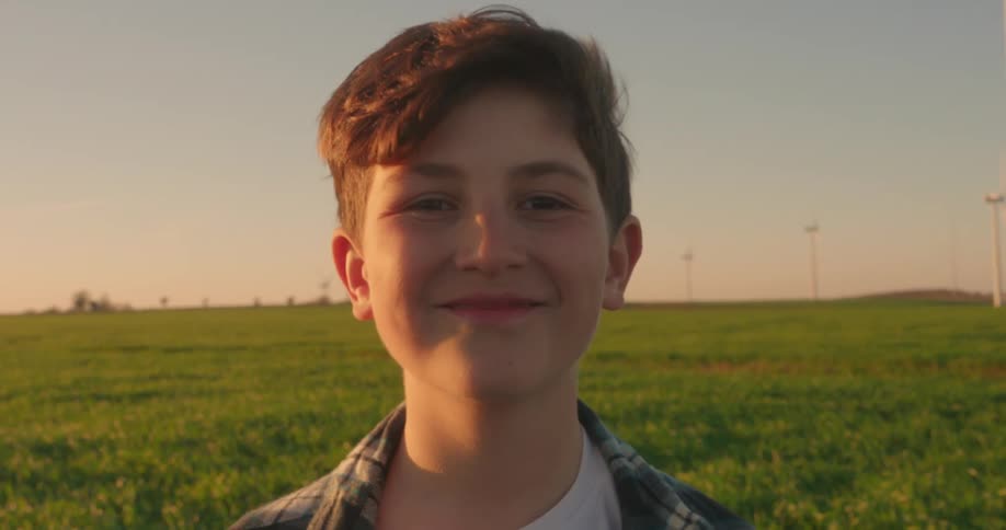 A boy stands in a green field with several wind turbines scattered across the landscape under a clear blue sky