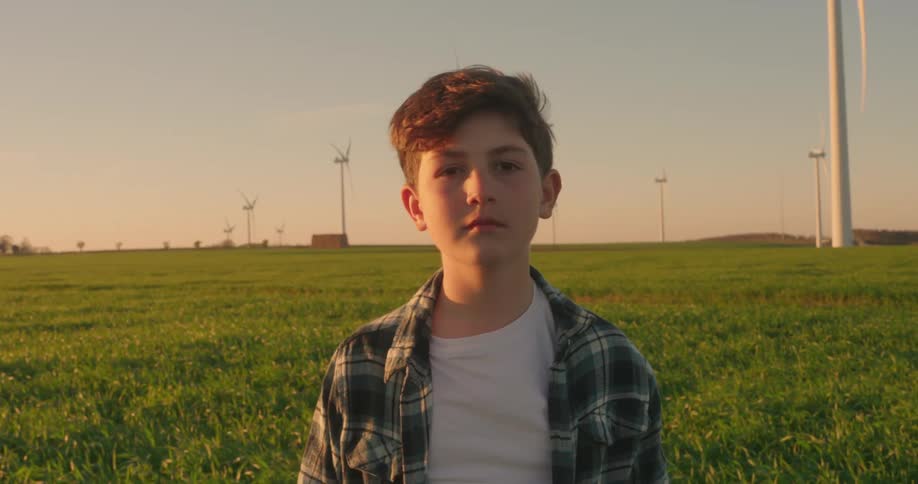 A young person with short hair stands in a field of tall grass gazing towards a row of wind turbines under a warm sunset sky