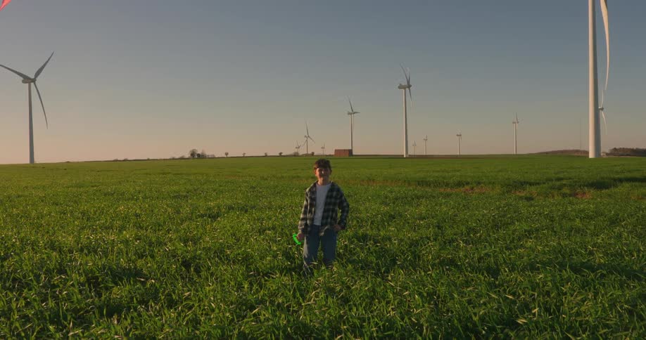A young person stands in a grassy field gazing up at a kite soaring against a backdrop of wind turbines under a clear sky