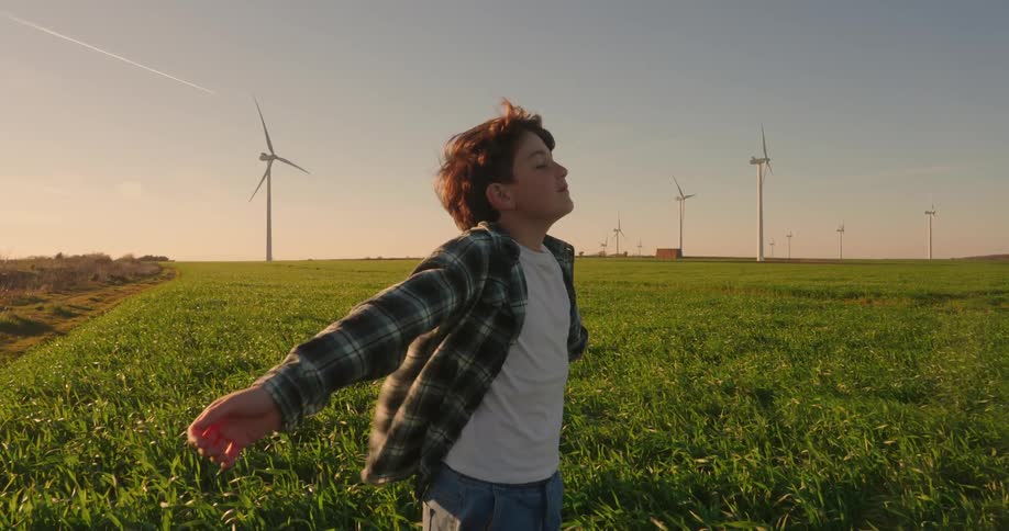 With his arms outstretched a young boy appears to be joyfully experiencing the breeze blowing through a vibrant green field alongside towering wind turbines