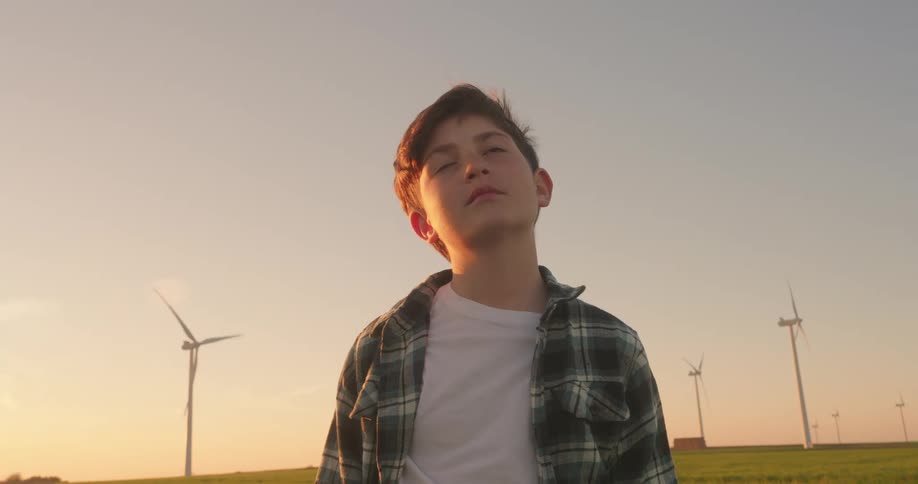 A young boy gazes upward at a field of towering wind turbines bathed in the warm glow of sunset