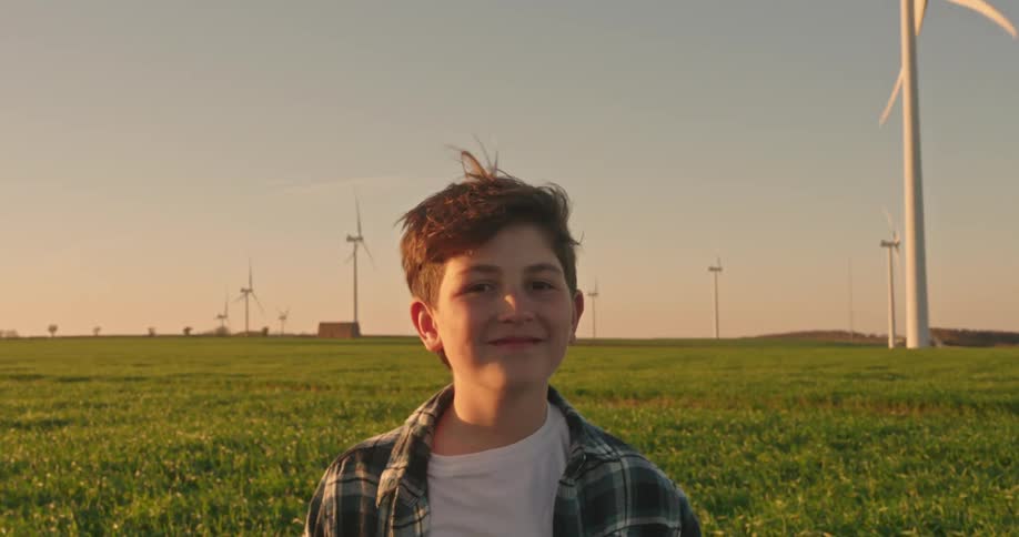 A young person with short hair smiles confidently while standing in a grassy field with wind turbines in the background under a warm sunset glow