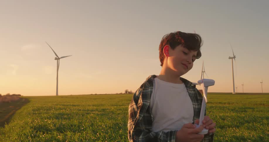 Boy holds a miniature wind turbine as he watches the toy spin in a field at sunset surrounded by real turbines