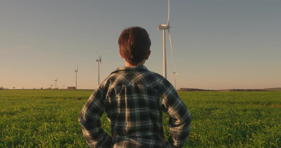 View of a boy from behind in an agricultural field watching wind energy platforms