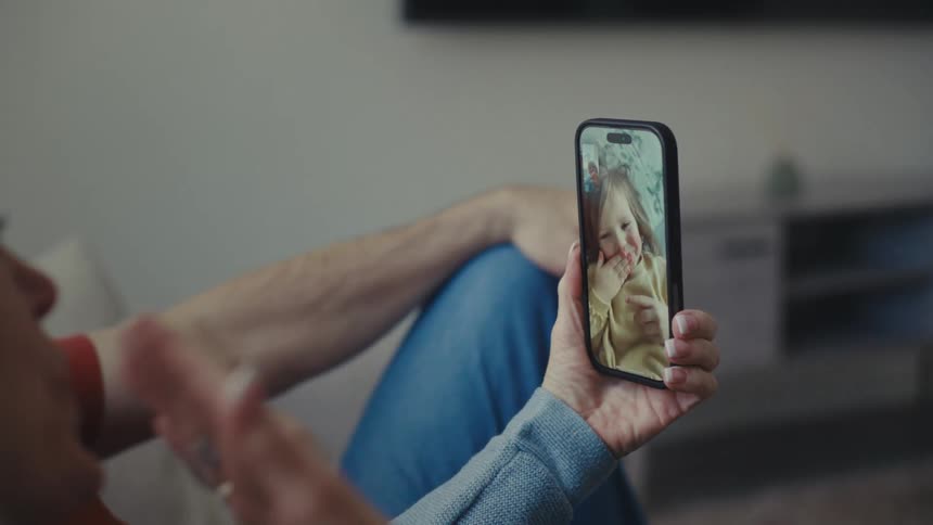 A man and woman are sitting on a couch while the woman holds a phone and blows kisses to someone on a video call