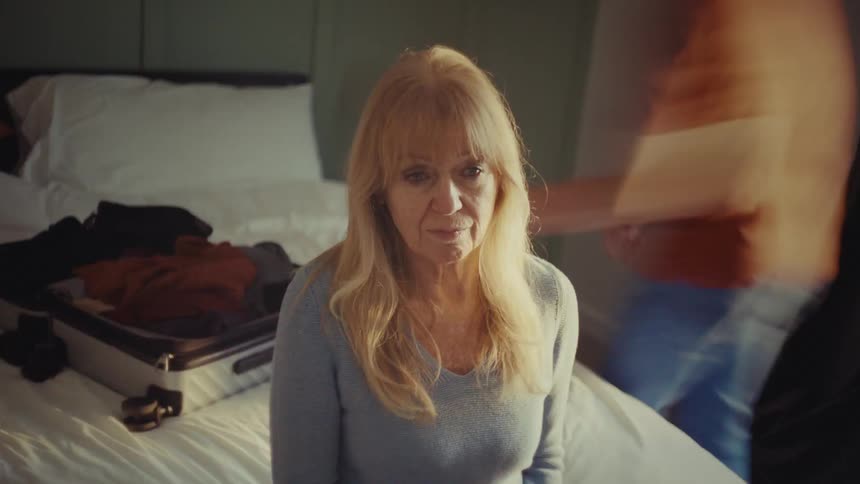 A thoughtful woman sits quietly on a bed as another person organizes clothes into a suitcase in a welllit room