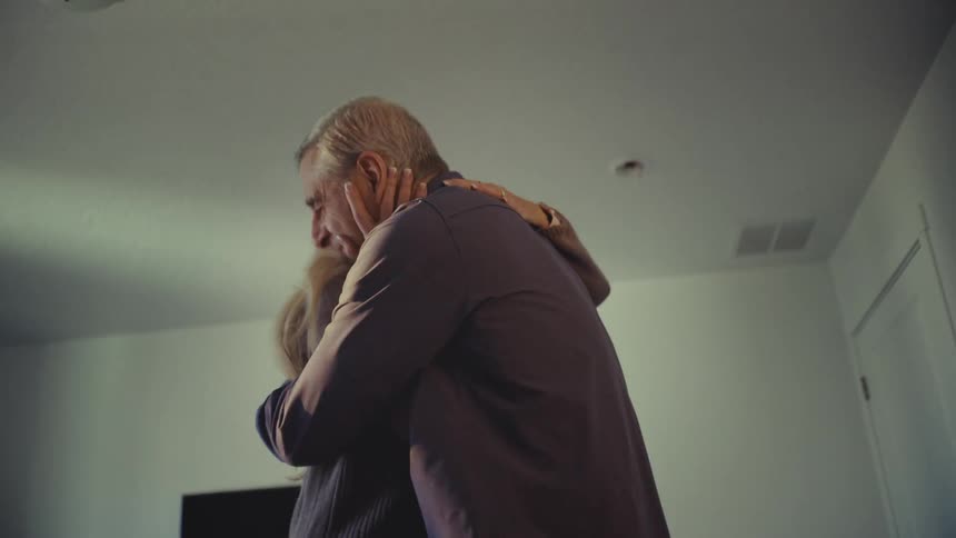 A man folds clothes in a bright sunlit bedroom as his wife hugs and kisses him affectionately