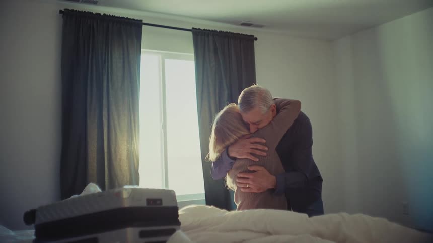 An older couple shares a tender embrace in a sunlit bedroom with a suitcase resting on the bed nearby
