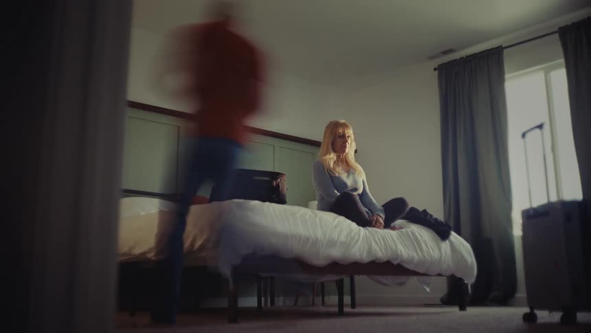 Smiling woman sits on the bed while her spouse packs a suitcase