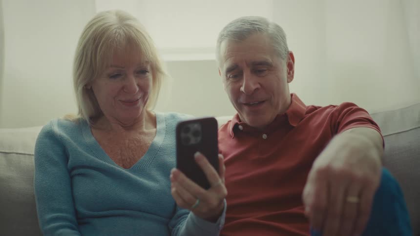 A man and a woman with gray hair are sitting on a white sofa talking on a video call using their cell phone which is in the womans hand and they are happy