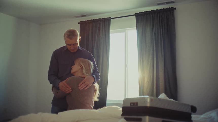 A standing man is hugging a woman who is sitting on a bed in an indoor setting with white walls