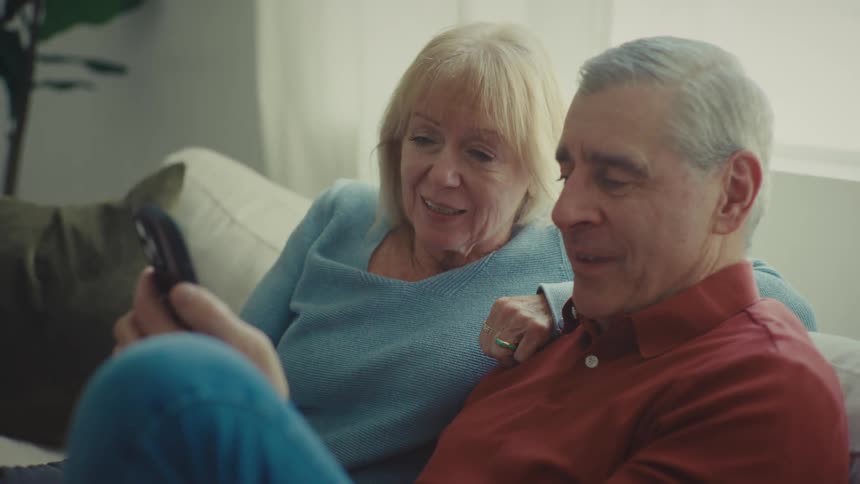 A couple sits on a couch in a bright room near a window smiling while engaged in a video call