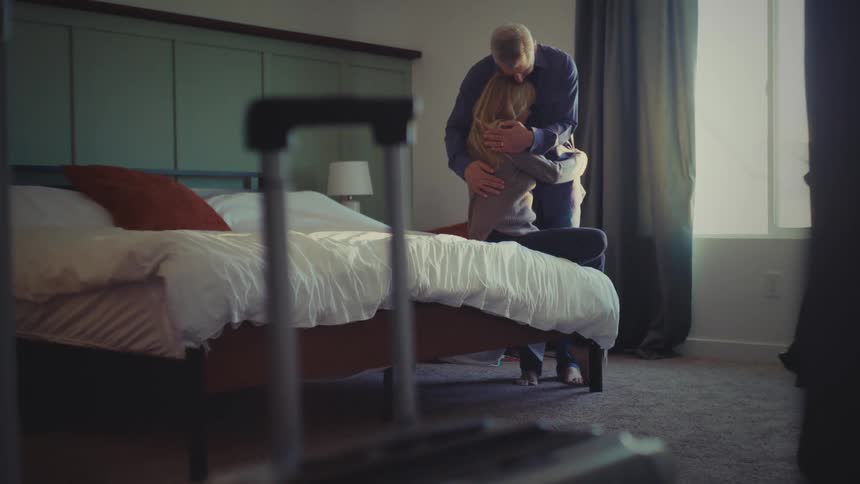 Through a slightly ajar doorway a couple embraces near a window in a hotel room with a suitcase prominently placed in the foreground