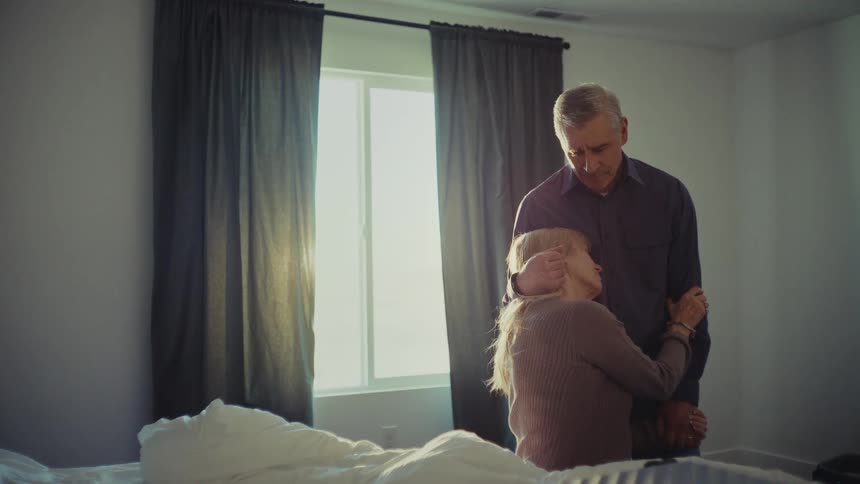 A woman sits on a bed while a man stands beside her embracing her in a sunlit bedroom with a curtaincovered window