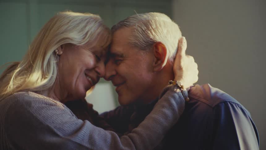 A mature couple shares an intimate and tender moment as a woman with long blonde hair gently holds the head of an older man with short gray hair