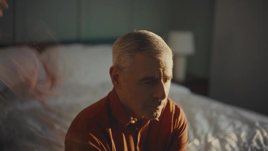 A man sits calmly on a bed as another person moves around him engaged in a heated argument in a brightly lit room