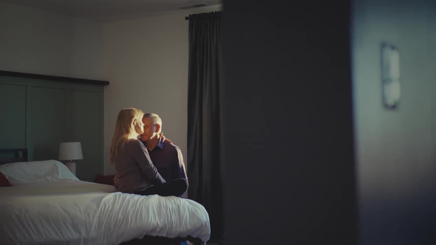 A woman sits on a bed while a man kneels beside her embracing her in a sunlit bedroom with a curtaincovered window