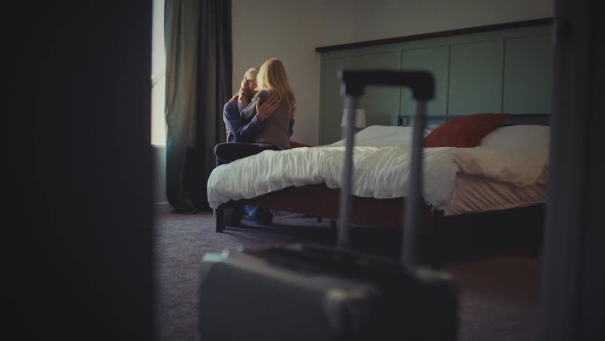 A couple hug on the bed with luggage by the door