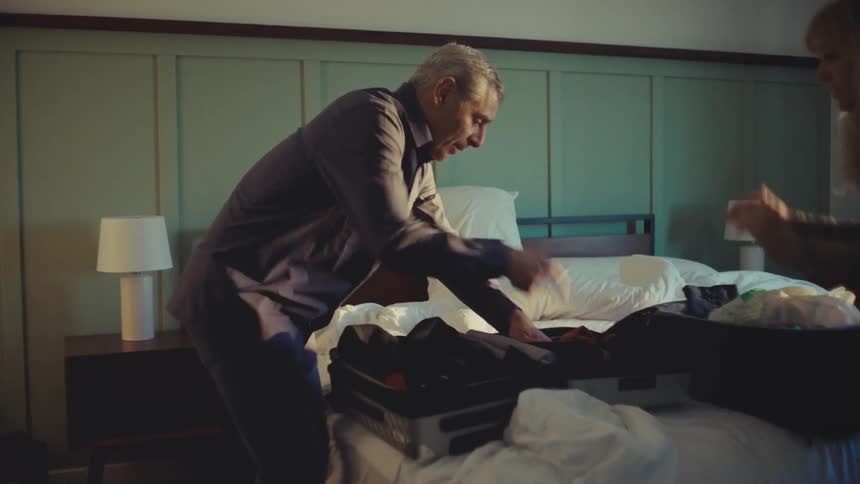 A couple hastily packs a couple suitcases laid open on the bed in a bedroom
