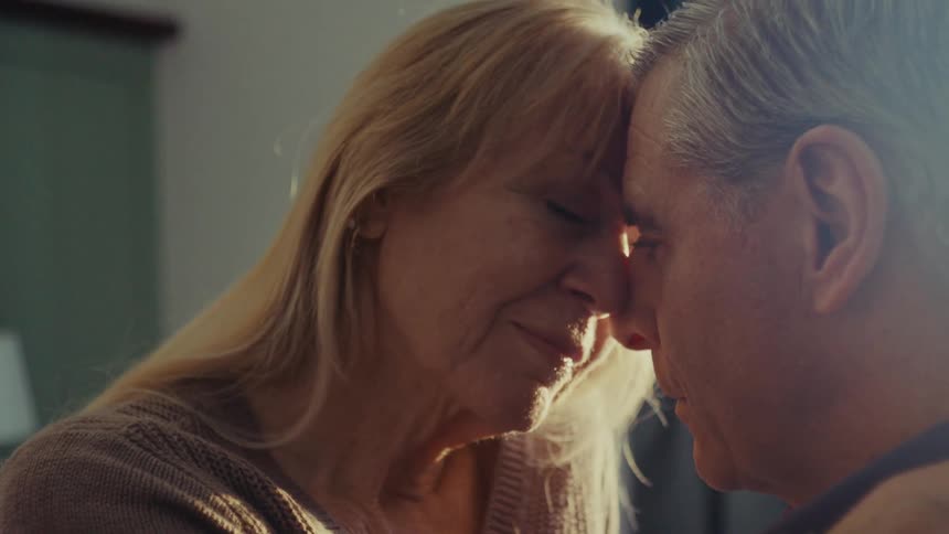 A mature couple shares an intimate and tender moment as a woman with long blonde hair gently holds the head of an older man with short gray hair