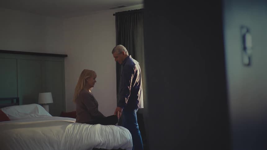 A woman sits on a bed while a man stands beside her embracing her in a sunlit bedroom with a curtaincovered window