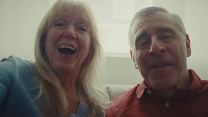 A couple sits on a couch in a bright room near a window smiling waving goodbye while engaged in a video call