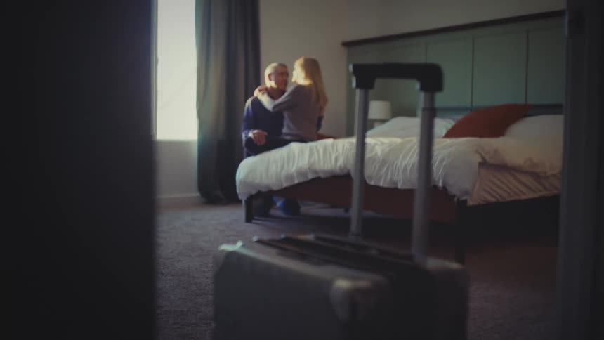 Through a slightly ajar doorway a couple embraces near a window in a hotel room with a suitcase prominently placed in the foreground