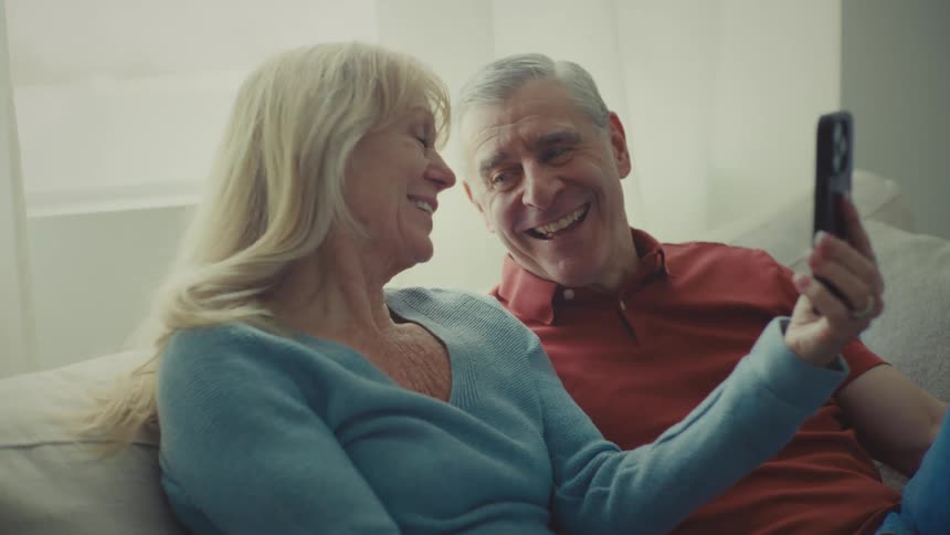 A couple sits on a couch one holding a smartphone while both are engaged in a video call