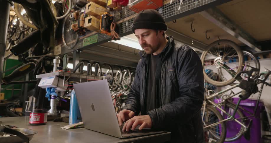 A man stands in a bicycle repair shop and types on a laptop computer